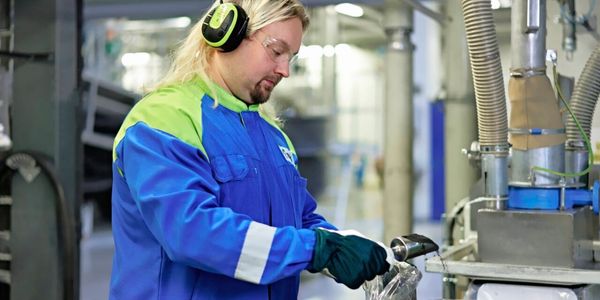 Factory worker wearing protective gear and headphones, adding black powder coating to a machine in an industrial setting.