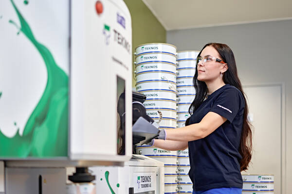 Teknos expert wearing safety glasses, operating a machine in a factory with stacked paint cans in the background.