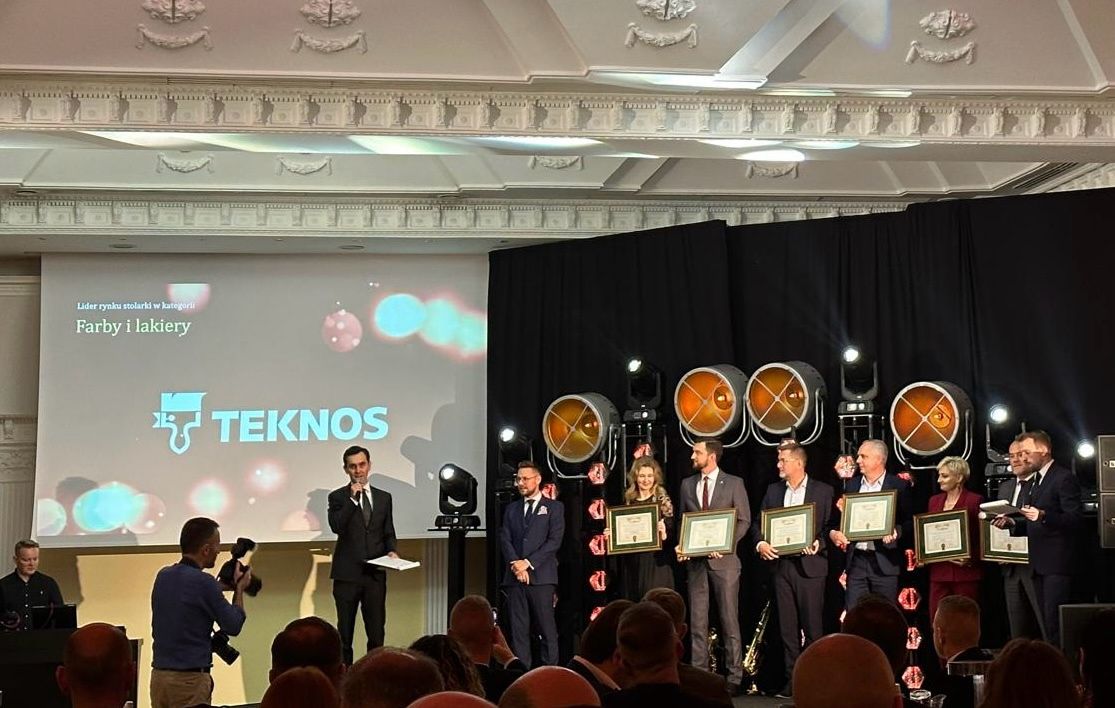 Group of award recipients standing on stage holding certificates at an awards ceremony, with Teknos logo displayed on a large screen in the background