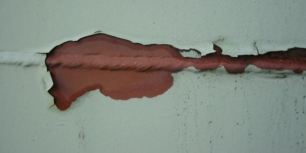 Close-up of a painted metal surface with peeling paint, exposing the underlying red primer and weld line.