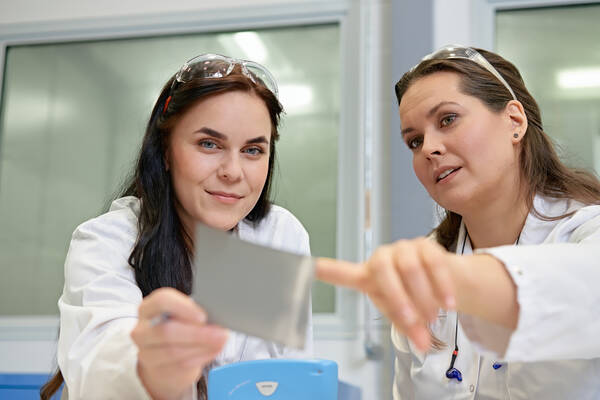 Two laboratory technicians in protective gear examining a metal sample, with one holding the sample and the other pointing, engaging in a discussion about its quality.